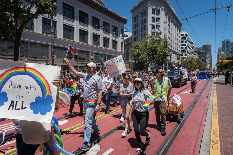 Julien, a bearded man wearing a clear hat, walks with a group of people holding signs featuring rainbow flags and the words 'Human rights for all' during San Francisco's Pride parade 2024. The group is walking on Market Street, with buildings visible in the background, shot from a side angle on a sunny day with a blue sky.