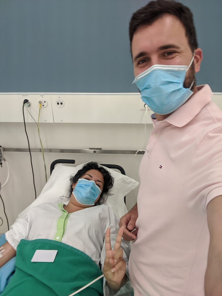 Marie-Rose, wearing a face mask, lies in bed before surgery for endometriosis, with her husband Julien, also wearing a face mask, standing at her bedside.