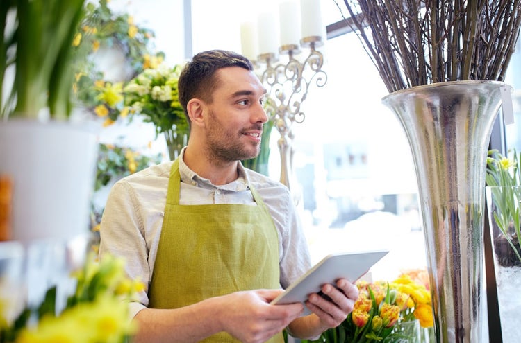 Portrait of a smiling male florist holding a tablet computer and looking away while working in a flower shop. He stands near vases filled with flowers, with the floral shop interior visible in the background.