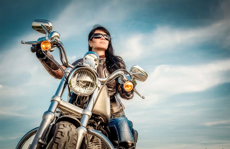 A woman sitting confidently on her motorcycle, wearing leather and sunglasses, holding the handlebars with both hands. Her long black hair flows, and she wears silver jewelry, adding a sense of mystery. The blue, cloudy sky in the background creates an atmosphere of freedom and adventure. Her focused gaze is directed ahead as she balances on the bike, capturing both her beauty and confidence in riding.