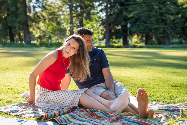 A happy couple sitting on a colorful blanket, laughing together as they pose for an engaging portrait. The man is wearing a navy blue short-sleeved shirt and shorts, while his wife wears a red top with a white long skirt. Both have brown hair and big smiles, conveying joy on a sunny day at the park