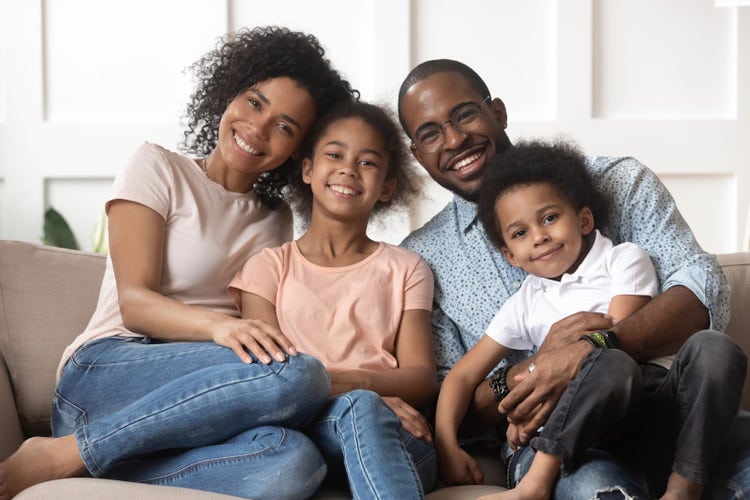 A happy Black family of four sitting on the couch in their living room, smiling and looking at the camera. The smiling parents are posing with their daughter and son for a family photo.