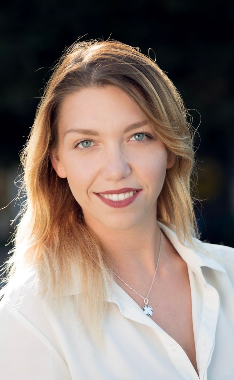 Professional portrait of a happy, attractive blonde woman in a white shirt, taken outdoors with a green lawn and a building in the background. This business headshot features natural lighting and high-resolution photography. Optimized for mobile view