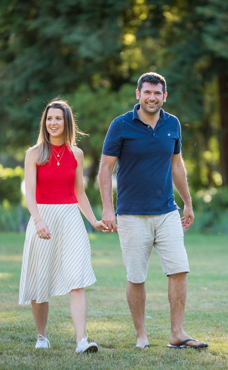 Full-body photo of an attractive couple walking hand in hand on the grass at San Mateo Central Park. He is wearing a blue polo shirt and shorts, and she is wearing a red top with a white skirt. The photo is taken during the golden hour on a summer day, with natural light and a background of green trees.