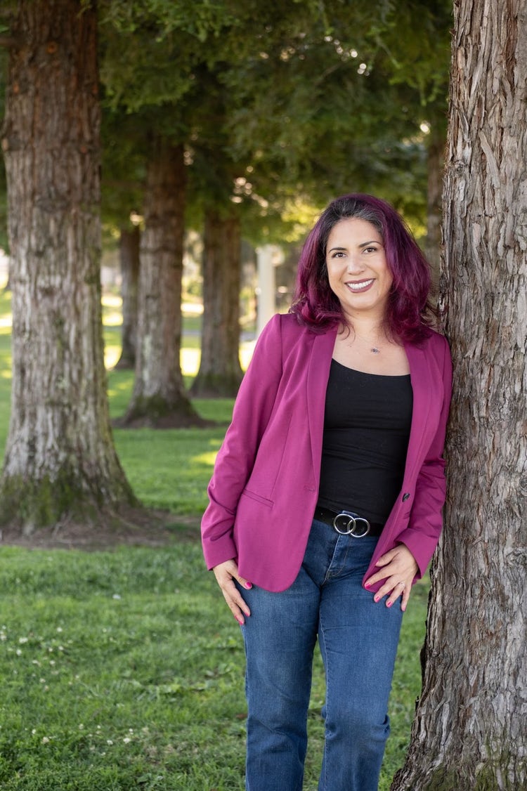 Full-body portrait of a round Latina woman with purple hair, wearing a blue and orange floral dress. She stands in a front yard with her left hand on her hip, showing tattoos on her left arm, and smiles at the camera. Green trees are visible behind her.