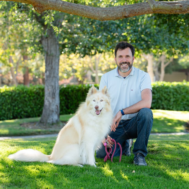 A professional photo of Julien, an attractive man in his early 40s wearing jeans and a light blue polo shirt, kneeling on the grass next to a cream-colored fluffy husky. Both have big smiles on their faces as they sit under a tree. The setting appears to be a park, as if they are out for a walk on a sunny day, with the shot taken at eye level.