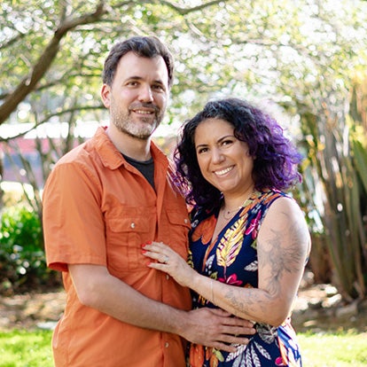A happy couple in their thirties posing for family photos outdoors on a sunny day. The tall, handsome man with short hair, Julien, is wearing an orange shirt and stands next to his wife, who has purple, curly, shoulder-length hair and tattoos on her left arm.