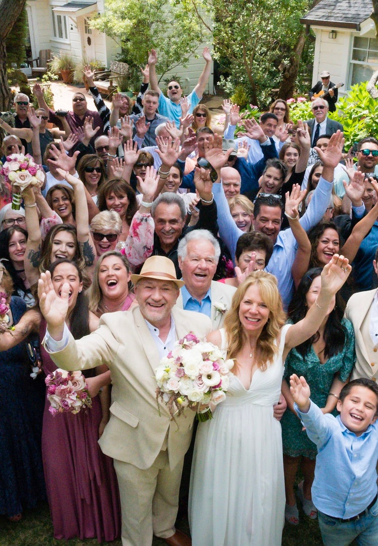 A photo of the entire wedding party celebrating in the front yard, with everyone waving and smiling at the camera. The bride stands at the front of the group next to the groom, who is wearing a beige suit. The scene includes a modern garden house in the background, and the photo is shot from above at Hastings House Garden Weddings. Optimized for mobile view