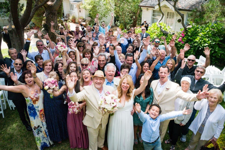 A photo of the entire wedding party celebrating in the front yard, with everyone waving and smiling at the camera. The bride stands at the front of the group next to the groom, who is wearing a beige suit. The scene includes a modern garden house in the background, and the photo is shot from above at Hastings House Garden Weddings