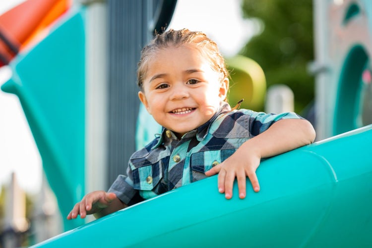 A young Hispanic boy wearing a blue plaid shirt smiles and looks at the camera while sitting at the top of a slide at a playground