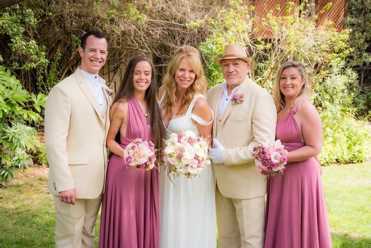 A wedding photo taken at Hastings House Garden Weddings, featuring the bride, groom, and their wedding party. The bridesmaids are in pastel pink dresses, and the groomsmen are in tan suits. From left to right: a man with short brown curly hair smiles at the camera, followed by a young woman with long brown hair in a pastel dress. The bride, with long blond hair and wearing a white dress, stands beside her husband, who is wearing a tan suit and a hat. On the far right is a young woman with blond hair in a pastel dress.